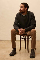 Man sitting on a wooden chair wearing a dark sweater, beige pants, and black shoes against a beige background.