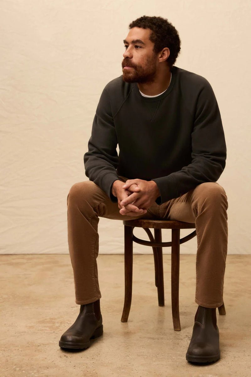 Man sitting on a wooden chair wearing a dark sweater, beige pants, and black shoes against a beige background.