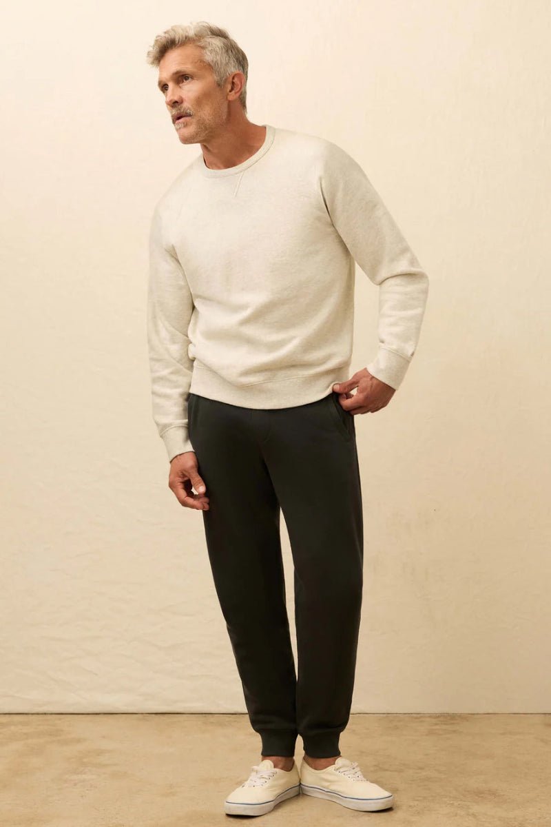 Man wearing a white sweater and black pants against a beige wall.