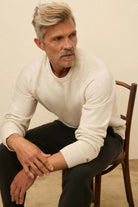 Man sitting on a wooden chair wearing a white sweater and black pants.