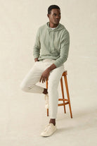 Man sitting on a wooden stool wearing a light green hoodie and white pants against a beige background