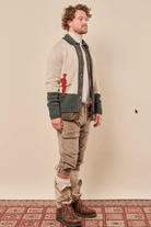 A man stands in profile wearing a cream knit cardigan with dark green trim and a red embroidered figure, layered over a white shirt and brown tie. He wears tan cargo pants, white gaiters, and brown lace-up boots against a plain beige wall and patterned rug.