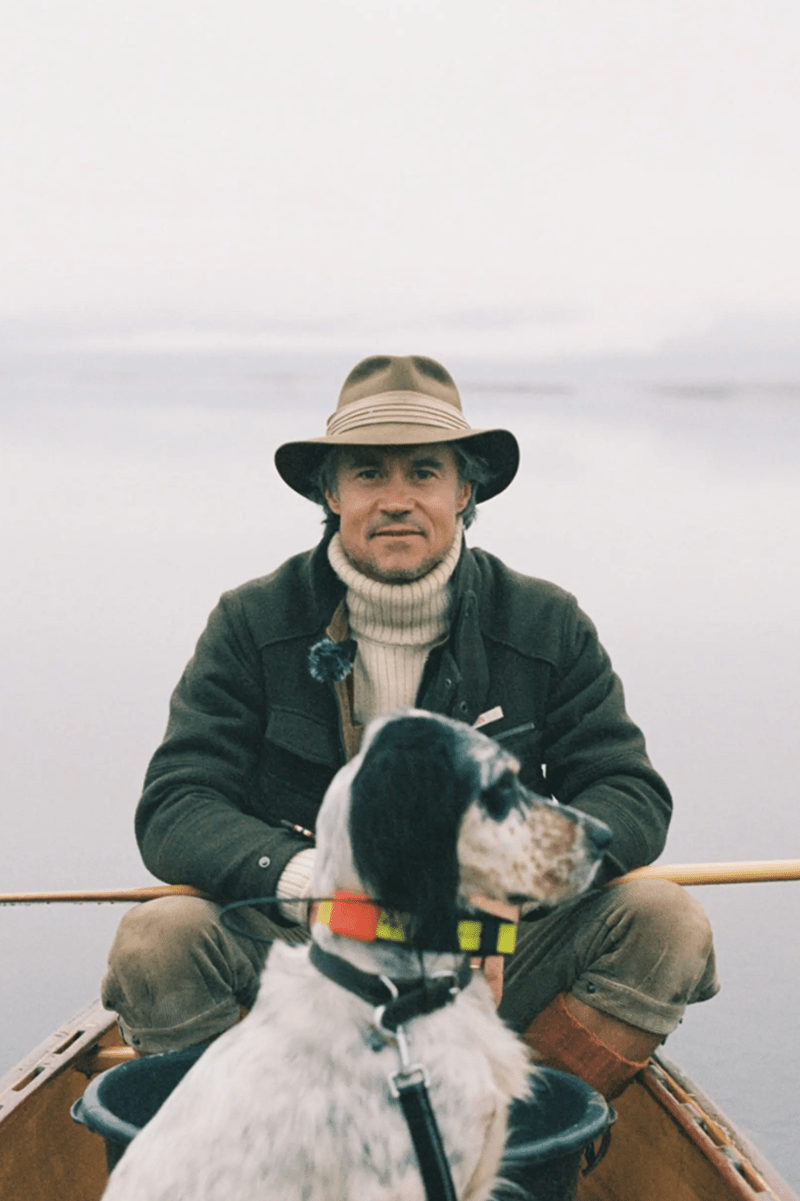 A man wearing a wide-brimmed hat, dark jacket, and cream turtleneck sits in a wooden boat with a black-and-white dog on a lake under an overcast sky.