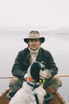 A man wearing a wide-brimmed hat, dark jacket, and cream turtleneck sits in a wooden boat with a black-and-white dog on a lake under an overcast sky.