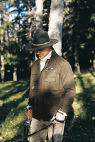 A man wearing a brown wide-brim hat with a yellow band, a brown wool-like jacket, and tan pants stands in a sunlit forest clearing holding a rifle.