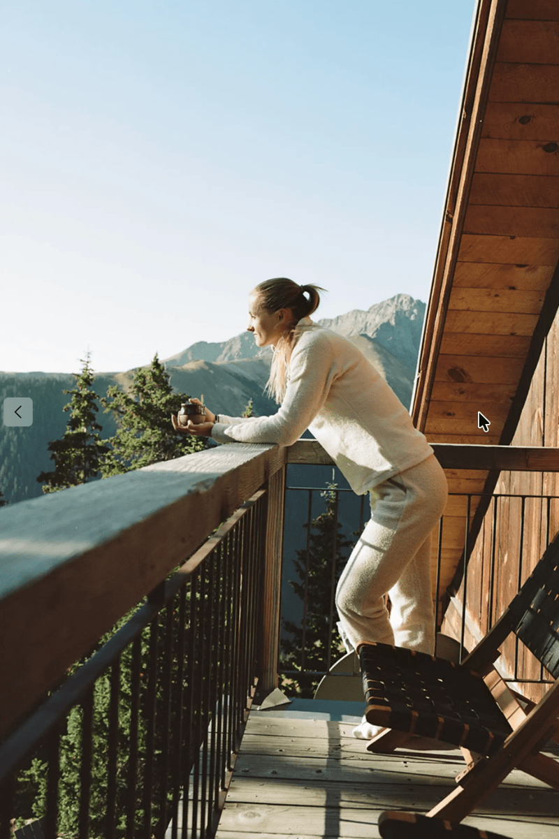 A woman in a cream-colored knit lounge set stands on a wooden deck of a cabin, looking out over a mountain and pine forest landscape.