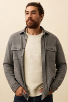 Man wearing a gray button-up shirt over a white sweater against a beige background