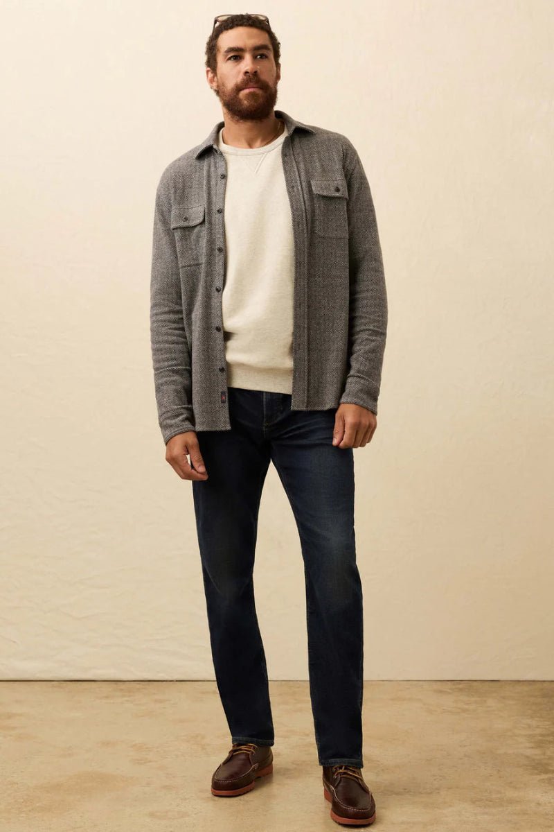 Man wearing a gray checkered shirt over a white t-shirt, dark jeans, and brown shoes against a plain background.