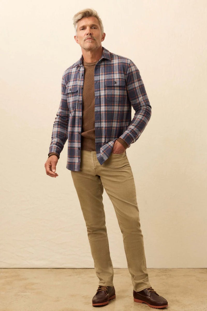 Man wearing a plaid shirt, brown sweater, beige pants, and brown shoes against a plain background