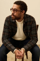 Man wearing glasses and a plaid shirt sitting on a chair against a beige background