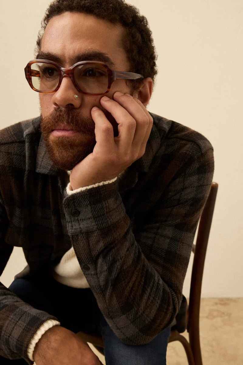 Man wearing glasses and a plaid shirt sitting on a chair against a beige wall.