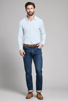 Man wearing a light blue shirt, dark blue jeans, and brown shoes on a gray background