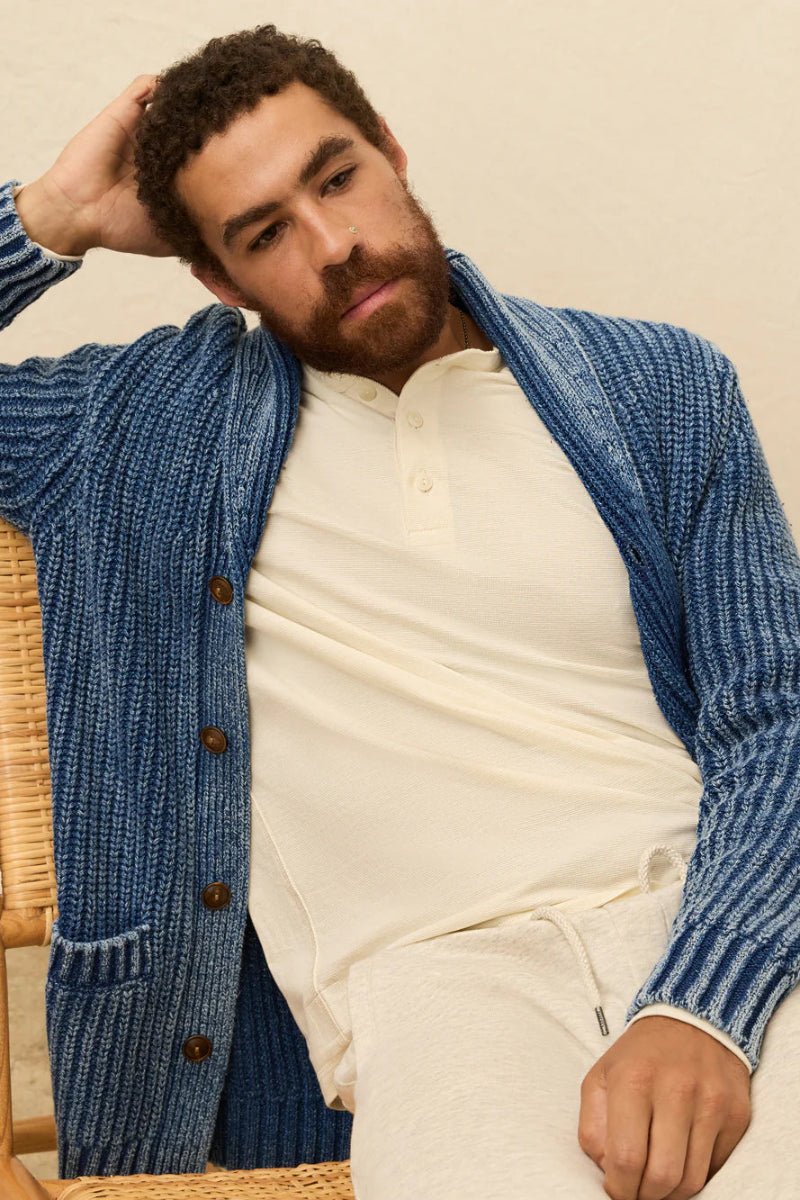 Man wearing a blue knitted cardigan over a white shirt, sitting on a wicker chair.