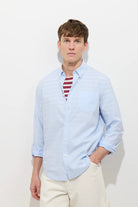 Man wearing a light blue shirt over a striped red and white shirt with beige pants on a white background