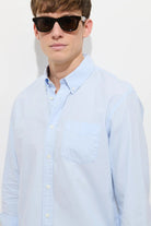 Man wearing a light blue button-up shirt and sunglasses against a white background