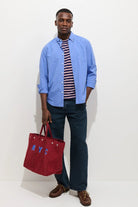Man holding a red bag with 'NYC' on it, wearing a blue shirt, striped shirt, and dark pants.