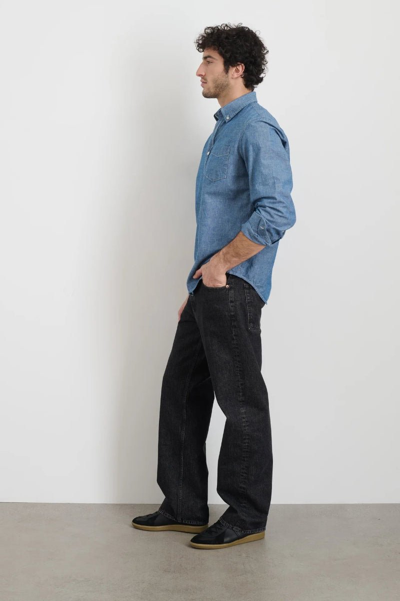 Man wearing a blue shirt and black pants standing against a white wall.