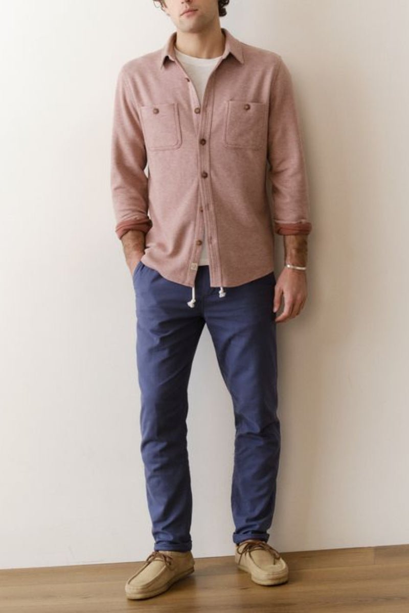 Man wearing a pink shirt, blue pants, and beige shoes against a white wall.