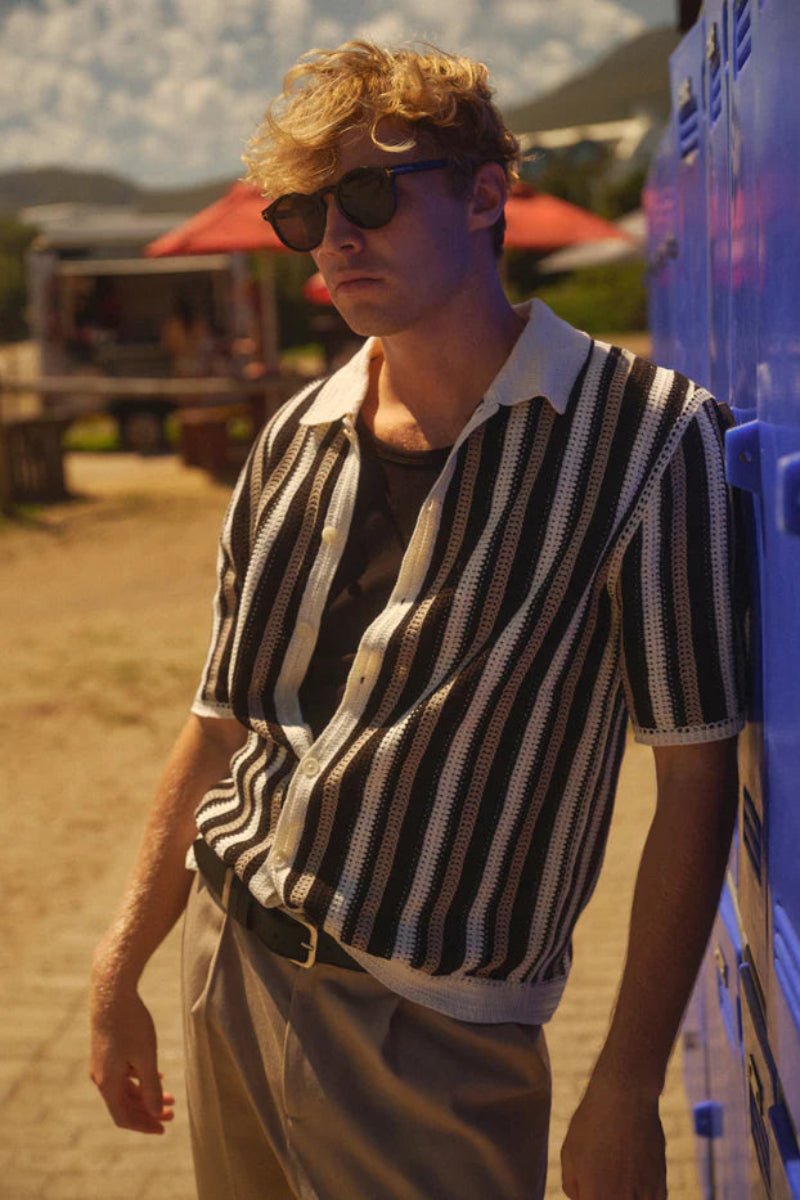 Man in a striped shirt and sunglasses standing next to a blue cooler outdoors.