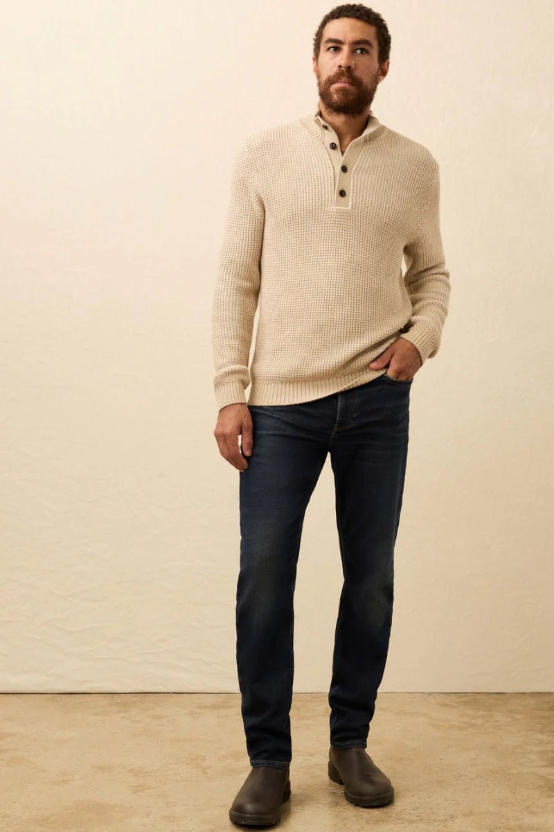 Man wearing a beige sweater and dark jeans against a plain background
