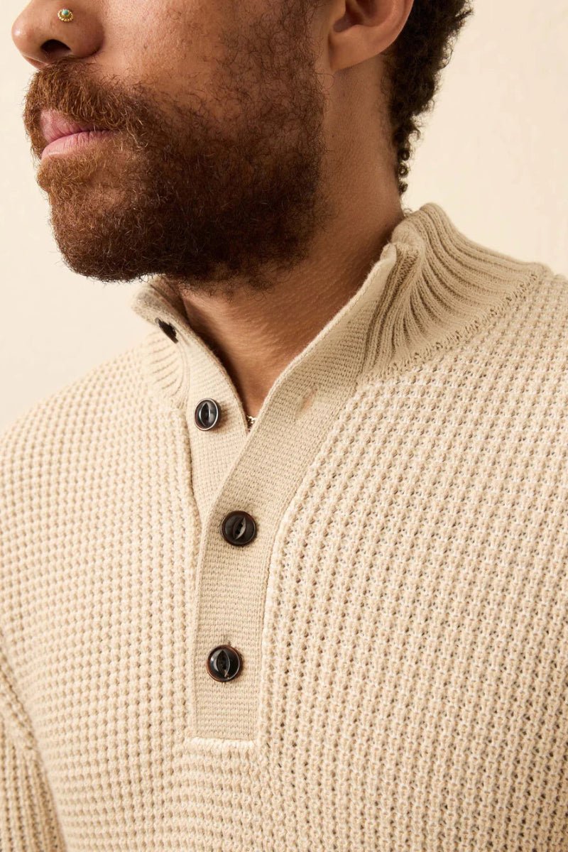 Close-up of a person wearing a beige knitted sweater with black buttons on a neutral background