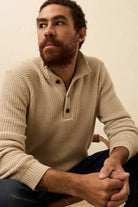 Man wearing a beige knitted sweater sitting on a chair against a plain background