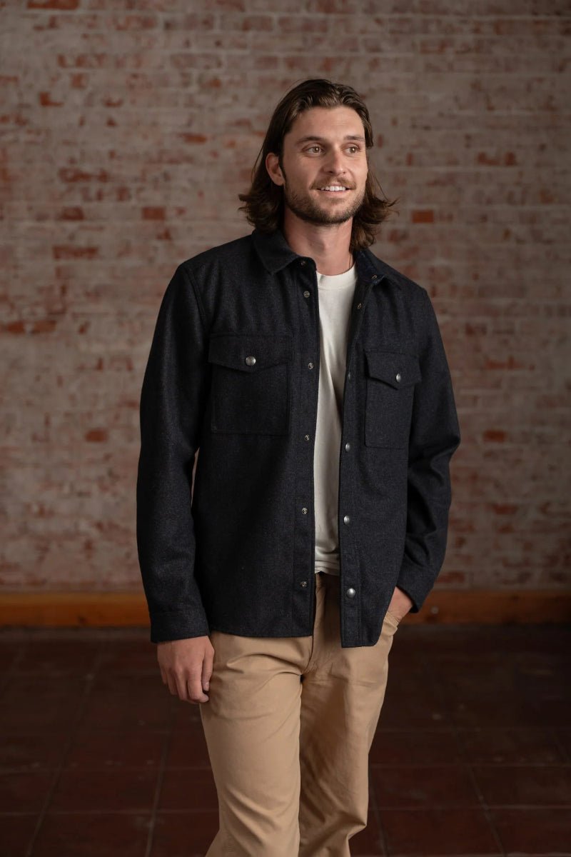 Man wearing a black jacket over a white shirt and beige pants against a brick wall.