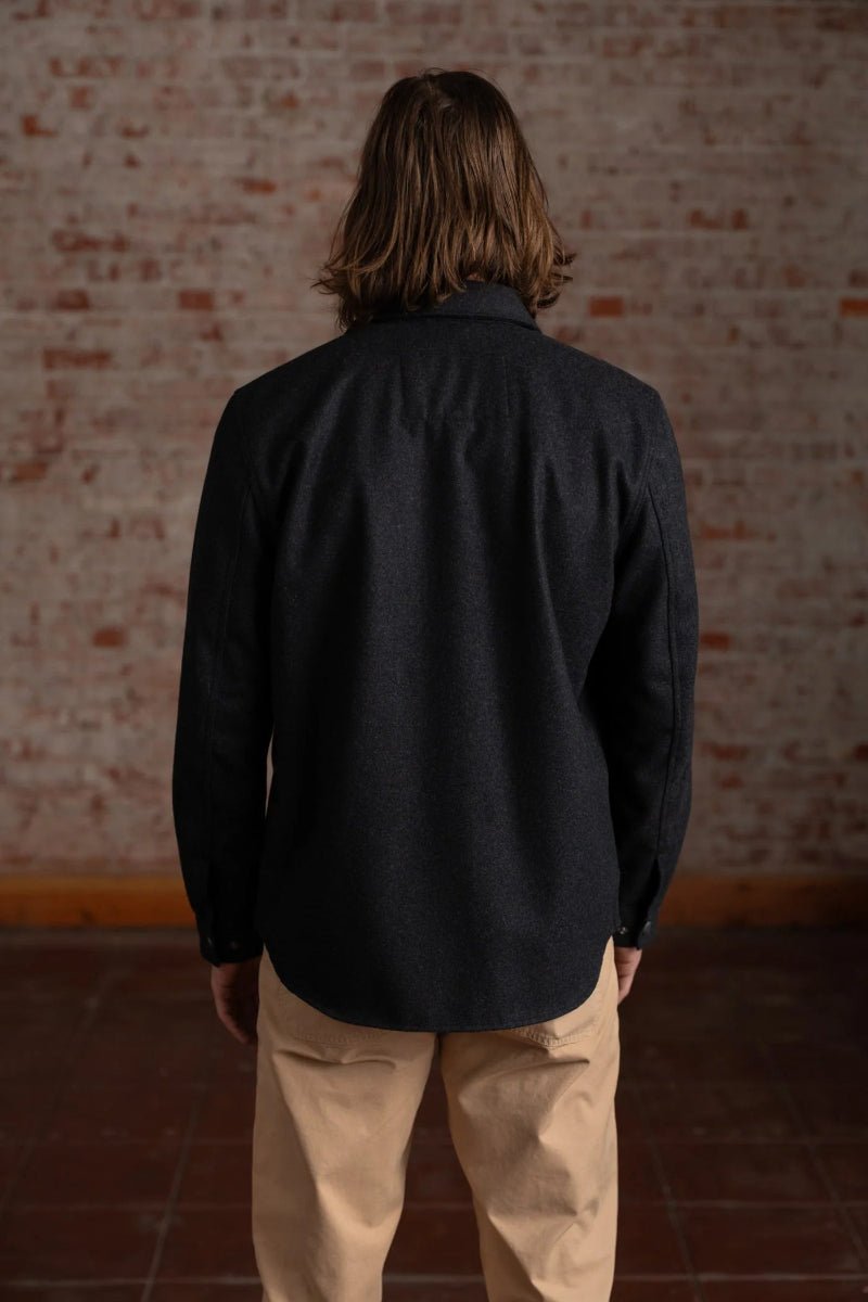 Person wearing a black jacket and beige pants standing against a brick wall.