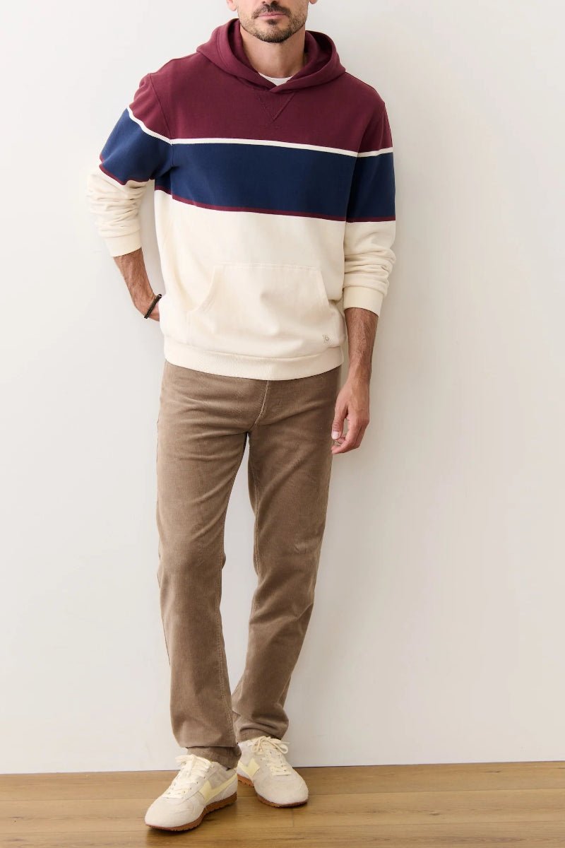 Person wearing a color-blocked hoodie and brown pants against a white wall.