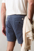 Person wearing blue shorts and a white shirt, holding a beige jacket against a plain background