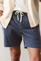 Person wearing blue shorts with a white shirt and beige jacket on a plain background