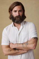 Man with a beard and tattoos wearing a white shirt against a beige background