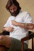 Man sitting on a chair wearing a white patterned shirt and green shorts, against a beige background.