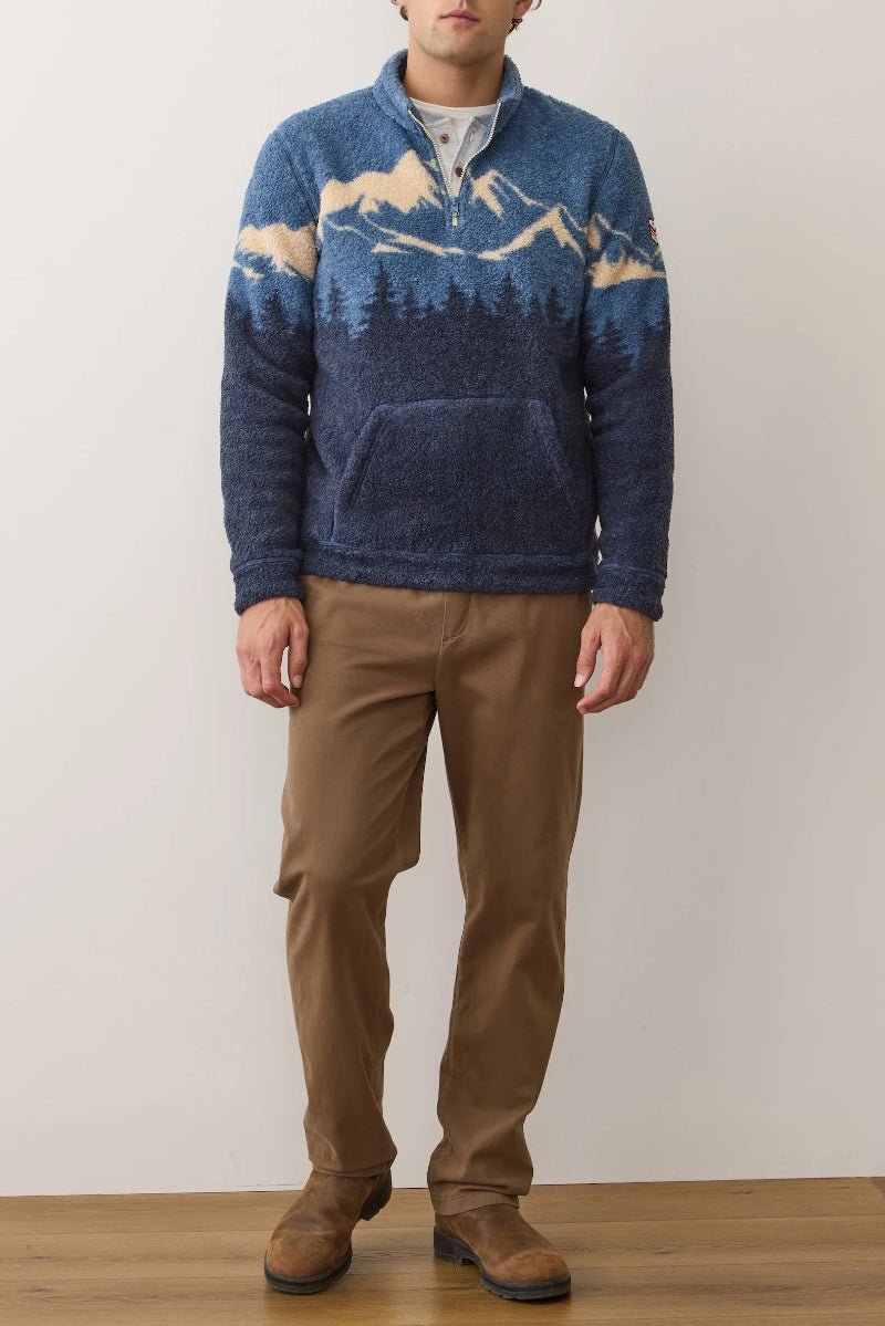 Person wearing a blue sweater with mountain design and brown pants against a plain background