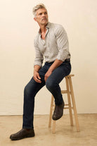 Man sitting on a wooden stool wearing a checkered shirt and dark jeans against a beige background