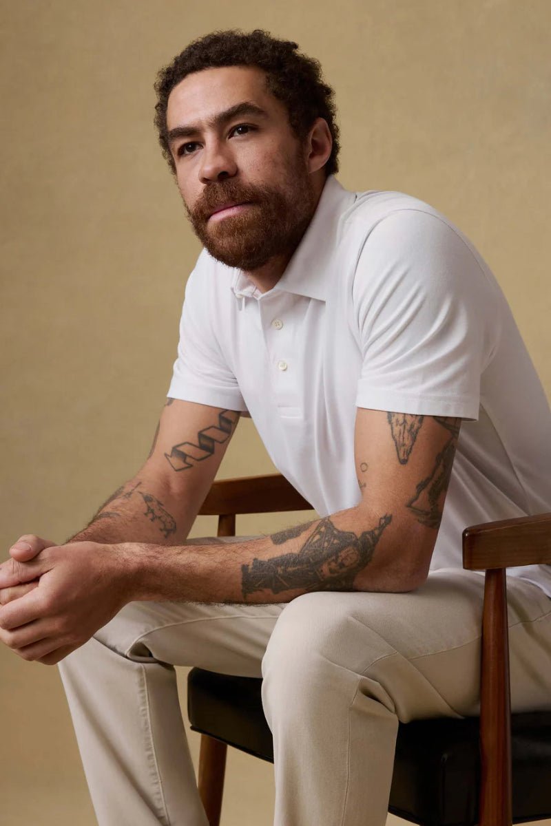 Man wearing a white polo shirt and beige pants sitting on a chair against a beige background