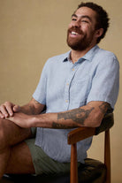 Man sitting on a chair wearing a light blue shirt and green shorts against a beige background