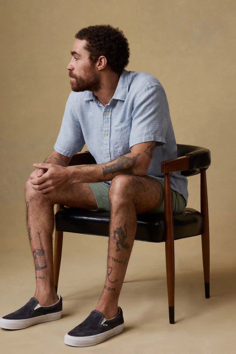 Man sitting on a chair wearing a light blue shirt and dark shoes against a beige background
