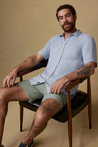 Man sitting on a chair wearing a light blue shirt and green shorts against a beige background