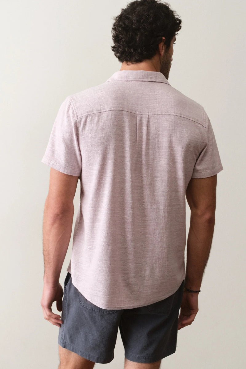 Man wearing a light pink short-sleeve shirt and dark shorts against a plain background