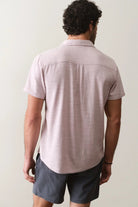 Man wearing a light pink short-sleeve shirt and dark shorts against a plain background