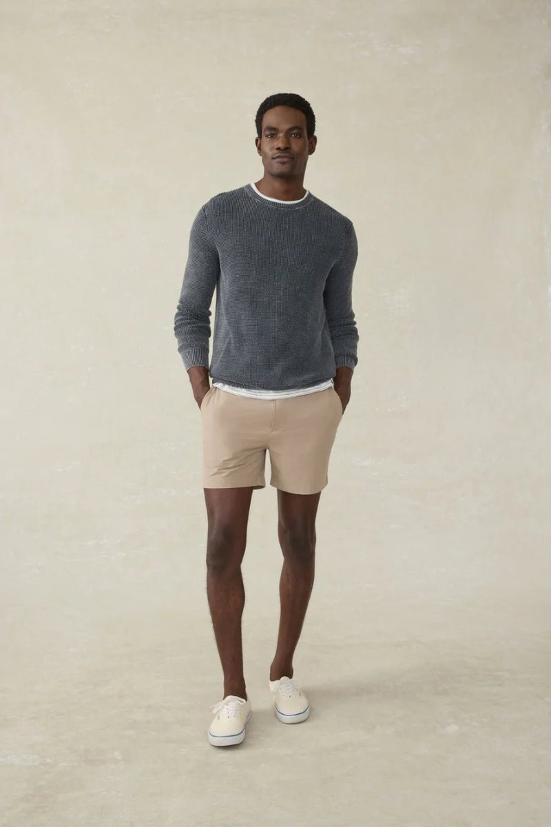 Man wearing a gray sweater, beige shorts, and white shoes on a plain background