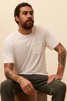 Man wearing a white t-shirt and dark jeans sitting on a wooden stool against a beige background