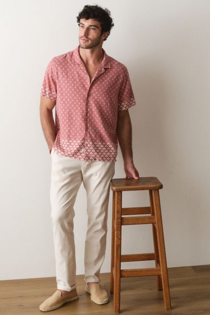 Man wearing a red patterned shirt and beige pants standing next to a wooden stool against a plain wall.
