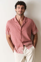 Man wearing a pink patterned shirt and white pants against a plain background