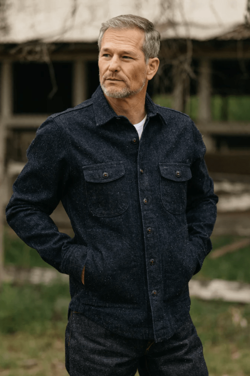Man wearing a dark denim jacket standing in front of a rustic building.