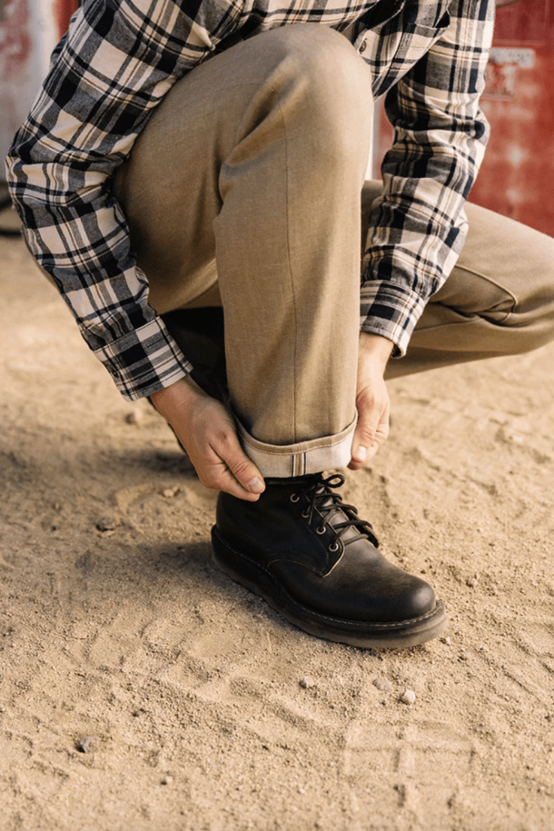 Person wearing a plaid shirt, beige pants, and black boots on a concrete surface.