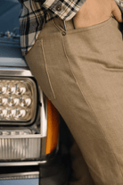 Person wearing brown pants leaning against a vehicle with a plaid shirt.