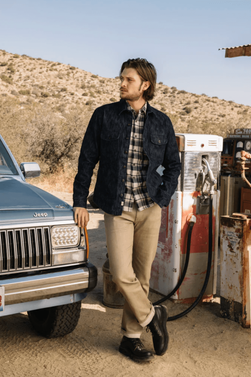 Man standing next to a vintage Jeep at a gas station with a desert landscape in the background