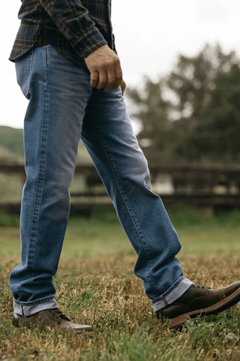 Person wearing blue jeans and brown shoes standing in a grassy field with trees in the background